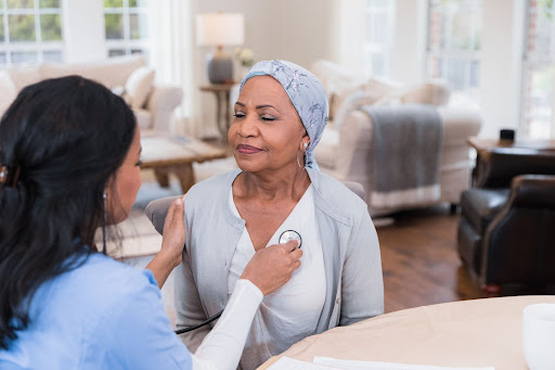 A home health aide in scrubs checks a female cancer patient’s heart with a stethoscope in the patient’s home