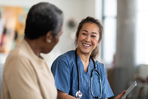 A skilled nurse wearing scrubs and stethoscope talks to an older person in their home